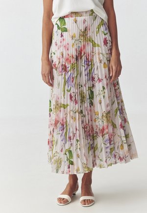 Femme portant une jupe midi blanche plissée avec un imprimé floral coloré, associée à des sandales blanches ouvertes à talons bas.