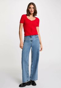 Top corto a maniche corte lavorato a maglia rosso con scollo a ondina e chiusura a bottoni, abbinato a jeans blu a gamba larga a vita alta e scarpe nere.