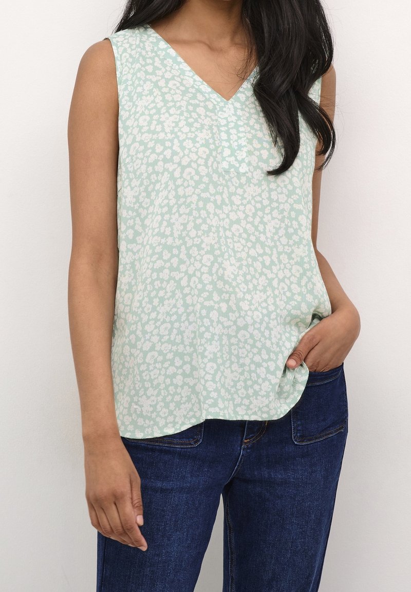 Femme portant un haut sans manches vert clair à motif floral blanc et un jean bleu foncé, main dans la poche, sur fond uni.