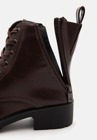 Botte marron à lacets avec un talon bloc bas, présentée avec une fermeture éclair latérale ouverte et une languette à l'arrière pour un enfilage facile.