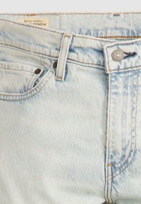 Section de la ceinture d'un jean en denim bleu clair montrant un bouton en métal, une passants pour ceinture, une poche avant et une étiquette à l'intérieur de la ceinture.