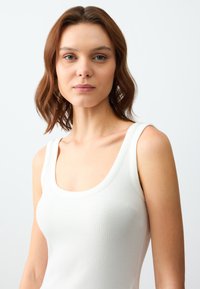 Top in cotone bianco a coste con scollatura ampia e spalline leggermente più spesse. Tessuto liscio, design aderente e senza motivi visibili.