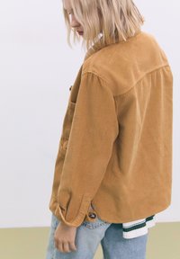 Giacca di velluto a coste di colore giallo senape, con una vestibilità comoda, tasche frontali e maniche arrotolate. Abbinata a una camicia a strati sotto.