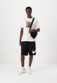 White t-shirt with colorful graphic, black shorts featuring white Nike logo, and white sneakers. Black crossbody bag includes zipper detail.