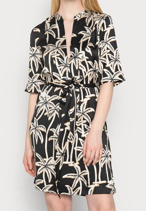 Czarna satynowa sukienka z beżowym nadrukiem palmy, krótkimi rękawami, dekoltem w serek i przewiązanym paskiem w talii, noszona przez stojącą kobietę.