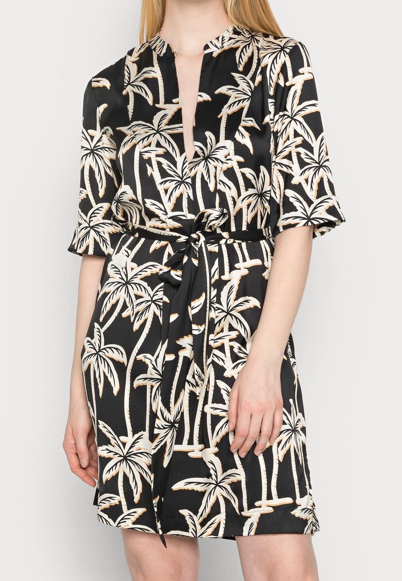 Robe en satin noir avec imprimé beige de palmier, manches courtes, col en V et ceinture nouée à la taille portée par une femme debout.