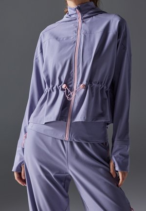 Mujer con chaqueta lila con cremallera y cintura con cordón ajustable y pantalones a juego, de pie frente a un fondo gris liso.