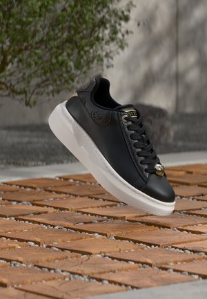 Baskets en cuir noir avec semelle blanche et lacets noirs, survolant des dalles en bois en extérieur avec un fond flou de verdure et un mur en béton.
