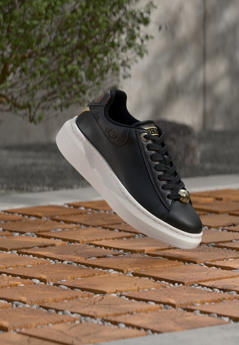 Baskets en cuir noir avec semelle blanche et lacets noirs, survolant des dalles en bois en extérieur avec un fond flou de verdure et un mur en béton.