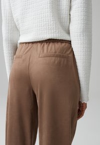 Brown suede-like pants with a relaxed fit, featuring back pockets. Paired with a textured white long-sleeve top.