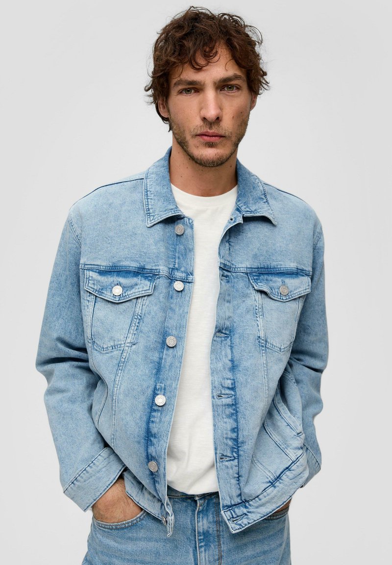 Jasnoniebieska kurtka dżinsowa zapinana na guziki, z dwiema kieszeniami na piersi i kołnierzem. Nosi się ją na białej koszulce z luźnym krojem.