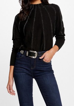 Kvinde iført en sort langærmet top med lodrette glitrende linjer, mørkeblå jeans og et sort bælte med en sølvfarvet udsmykket spænde.