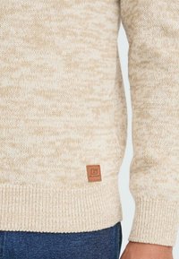 Beige gebreide sweater met geribbelde zoom en manchetten, gedragen met blauwe jeans; heeft een klein bruin leren label met de tekst "Blend" nabij de zoom.