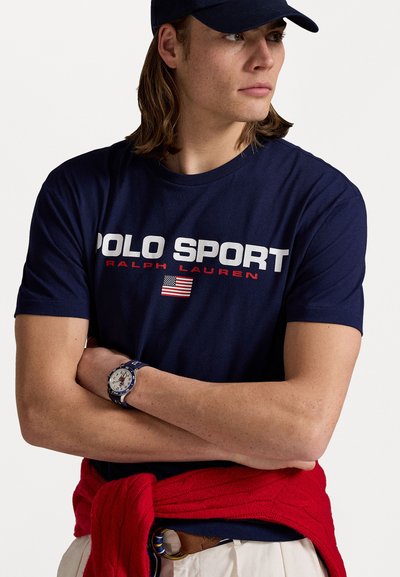 T-shirt en coton bleu marine avec le logo "POLO SPORT RALPH LAUREN" en blanc et rouge, présentant un drapeau américain. Pull en tricot rouge drapé autour de la taille.