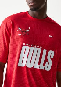 Κόκκινη φαρδιά μπλούζα διχτυωτού υφάσματος με την επιγραφή "CHICAGO BULLS" σε μεγάλα λευκά γράμματα. Διαθέτει λογότυπο με μαύρο και λευκό ταύρο με λεπτομέρειες. Στρογγυλή λαιμόκοψη.