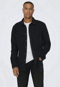 Chemise en denim noir avec des boutons à l'avant, deux poches poitrine et des manches longues. Portée par-dessus un T-shirt blanc, associée à un jean noir. Design simple.