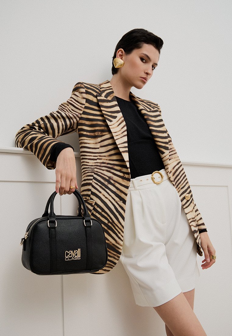 Sac à main en cuir noir avec un logo doré, doté de poignées arrondies. Le modèle porte un blazer rayé marron et un short blanc, posant devant un fond neutre.