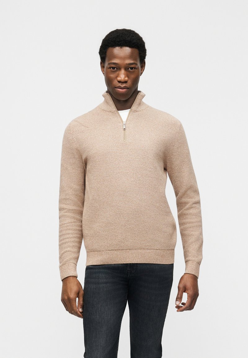 Νεαρός άνδρας που στέκεται στραμμένος προς τα εμπρός, φορώντας ένα μπεζ πουλόβερ με φερμουάρ στη μέση και σκούρα τζιν, σε απλό λευκό φόντο.