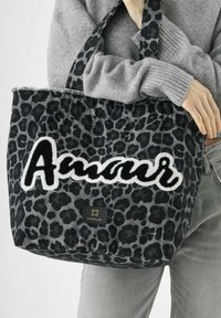 Personne portant un pull gris et un jean tenant un sac fourre-tout à imprimé léopard noir et gris avec le texte "Amour" et le logo Codello.