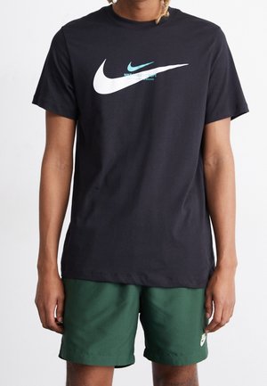 Schwarzes Baumwoll-T-Shirt mit großem weißem Nike Swoosh-Logo und einem kleineren blauen Akzentlogo, kombiniert mit grünen Shorts, glatte Textur.
