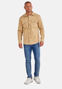 Beige Button-Up-Hemd mit zwei Brusttaschen, kombiniert mit blauen Jeans und weißen Sneakern. Das Hemd hat einen Kragen und einen lässigen Schnitt.