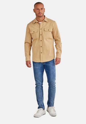 Beige button-up shirt with two chest pockets, paired with blue jeans and white sneakers. The shirt has a collar and a casual fit.