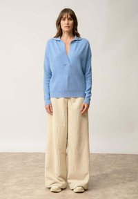 Jasnoniebieski sweter z dzianiny z dekoltem w serek i kołnierzykiem, połączony z beżowymi szerokimi spodniami z prążkowanej tkaniny i jasnym obuwiem.