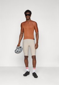 La tenue de cyclisme comprend un maillot marron ajusté à manches courtes, un short beige, des chaussures de cyclisme noires et un casque gris avec un design texturé.