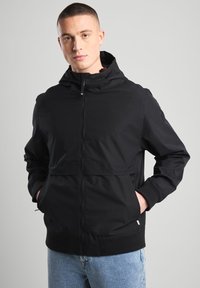 Black zip-up jacket with a hood, featuring a smooth texture and elastic cuffs. Designed with a front pocket and subtle branding on the sleeve.