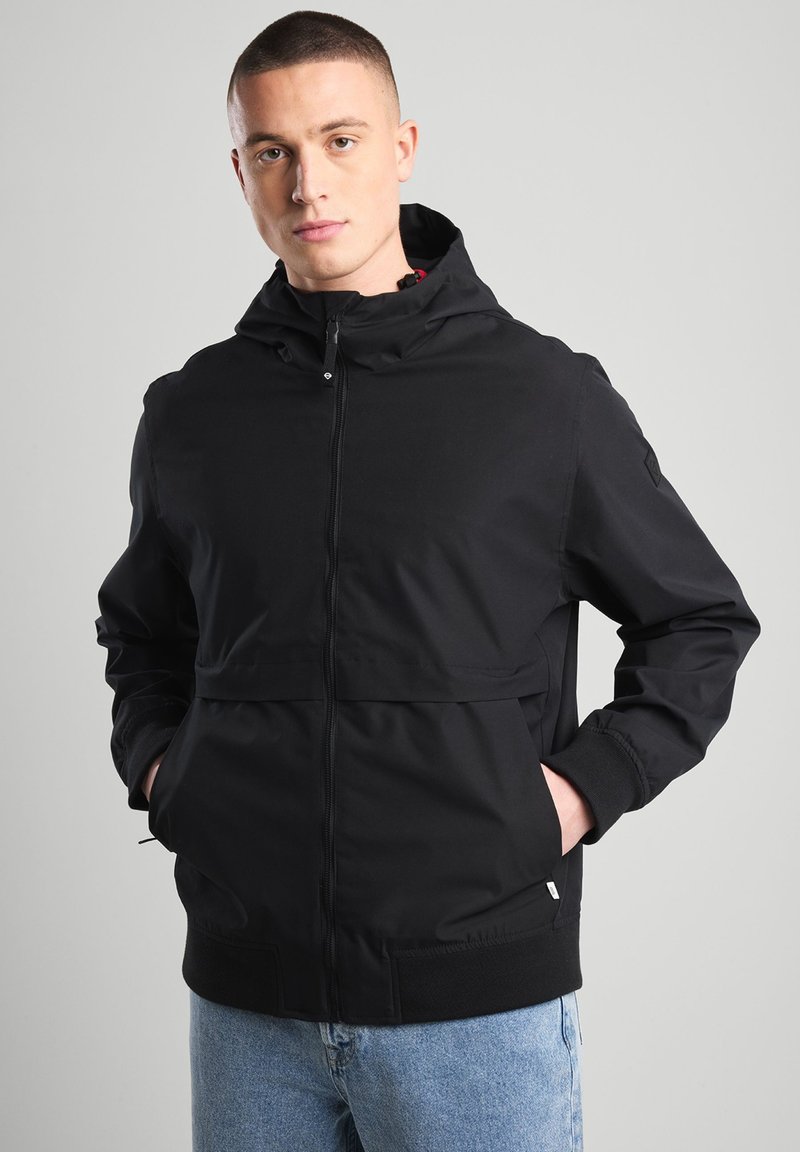 Black zip-up jacket with a hood, featuring a smooth texture and elastic cuffs. Designed with a front pocket and subtle branding on the sleeve.