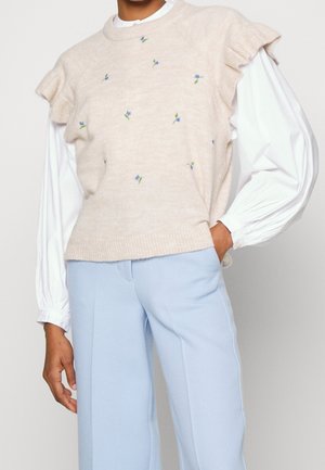 Pull en laine beige avec des fleurs bleues brodées et des accents verts, doté d'épaules à volants. Associé à un pantalon bleu clair.