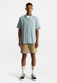 Lichtblauwe kortgebouwde blouse met een gestructureerde stof, gecombineerd met beige shorts, witte sokken met rode letters en zwarte sneakers.