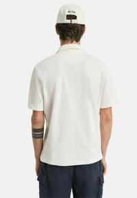 Weißes Poloshirt mit lockerer Passform und kurzen Ärmeln, versehen mit einem kleinen Bestickungsdetail am Rückenkragen. Glattes Material und schlichtes Design.