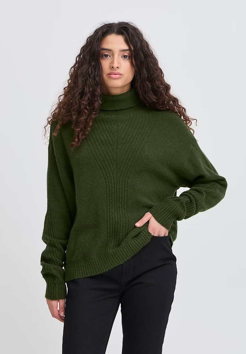 Maglione verde lavorato a maglia con collo alto, texture a coste e spalle basse, abbinato a pantaloni neri, su uno sfondo chiaro.