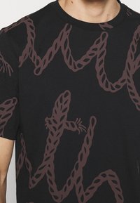 Černé tričko s hnědým vzorem připomínajícím provaz, do kterého jsou integrovány tvary ještěrek; kulatý výstřih, krátké rukávy, bavlněný materiál.