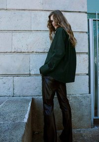 Oversized dark green shirt with long sleeves paired with shiny black leather pants. Textured stone background with a vertical window.
