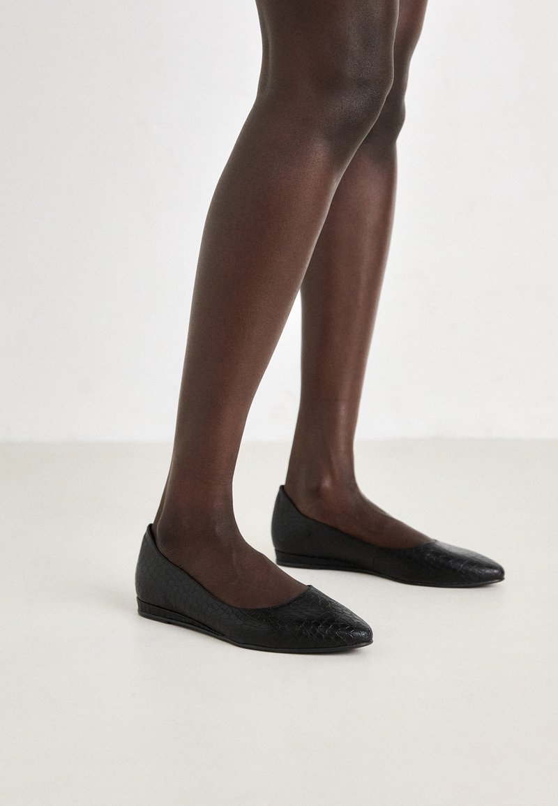 Schwarze flache Schuhe mit einem strukturierten, reptilienähnlichen Muster. Spitz zulaufende Zehen und niedrige, flexible Absätze. Getragen mit transparenten schwarzen Strumpfhosen.