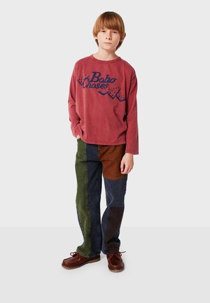 Red long-sleeve shirt with blue graphic text, paired with multi-colored patchwork pants in green, brown, and navy. Brown footwear completes the outfit.