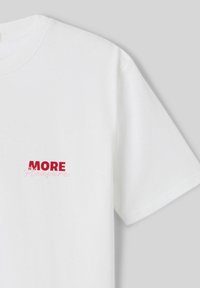 T-shirt en coton blanc avec des manches courtes et un col rond. Impression sur le devant avec le mot "MORE" en rouge vif et le mot "Pleasure" en rose doux.