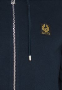 Marineblå hettejakke med s�ølvglidelås, snor med metallspiss, og en svart lapp med gulldetaljer som har Belstaff-logoen.