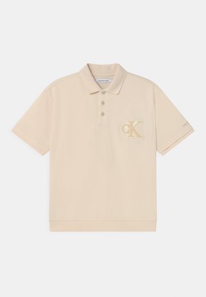 Polo beige en coton, doté d'une patte de boutonnage à trois boutons et d'un logo "CK" brodé sur la poitrine gauche. Texture douce.