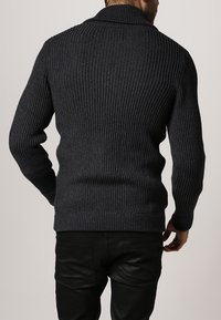 Mörkgrå ribbad tröja med en vikkrage och textilstruktur, bärs över ett par svarta byxor. Passformen är kroppsnära.