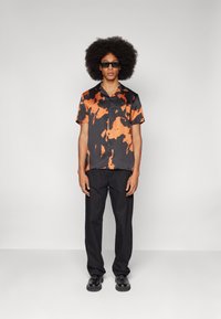 Korte mouwen zwarte shirt met oranje abstracte patronen, gecombineerd met zwarte broek en zwarte schoenen. Glad stof, relaxte pasvorm, met knopen ontwerp.