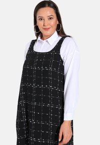 Robe longue à carreaux noirs avec un motif en grille blanche, décolleté carré et sans manches, fabriquée en tissu texturé, associée à une chemise blanche.