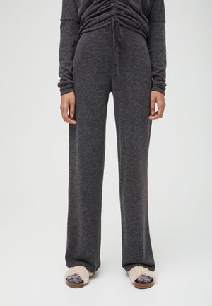 Woman wearing dark gray wide-leg pants and matching ruched top, paired with beige fuzzy open-toe slippers, standing against white background.