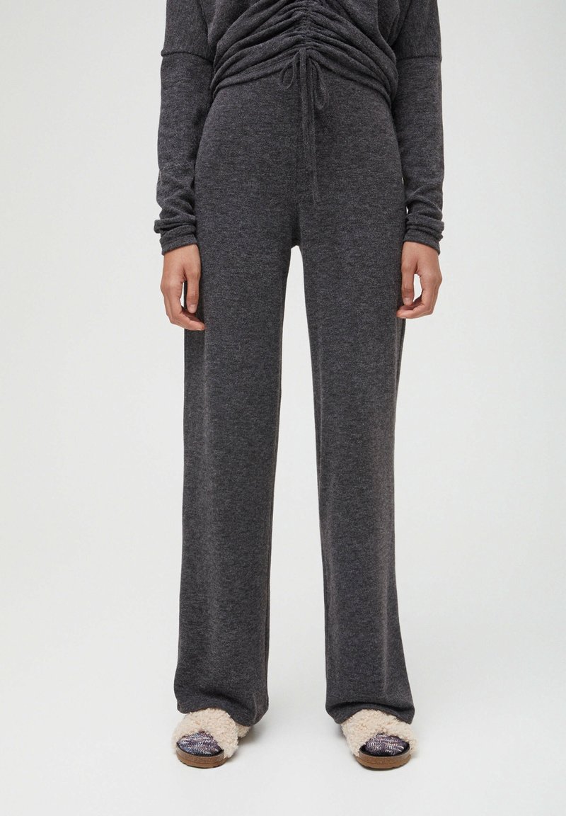 Femme portant un pantalon large gris foncé et un haut froncé assorti, associé à des chaussons beiges à bouts ouverts et poilus, debout devant un fond blanc.