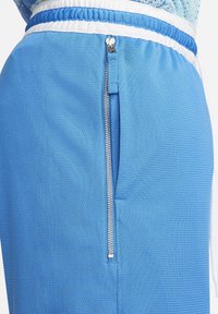 Gros plan sur un short texturé bleu avec une ceinture élastique blanche et bleue et une poche zippée verticale argentée sur le côté.