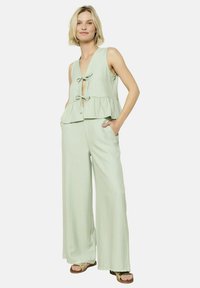 Svetlo zelen, brezrokaven vrh s kratkim V-izrezom in nabranim robom ter zavezovanjem, v kombinaciji s hlačami širokega kroja. Mehka, teksturirana tkanina z navpičnimi črtami.