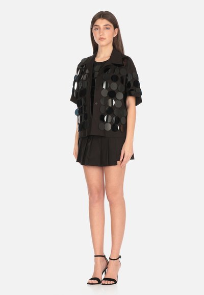 Camicia nera a maniche corte con paillettes circolari, abbinata a una mini gonna a pieghe nera e tacchi neri con cinturino, in piedi su uno sfondo bianco.