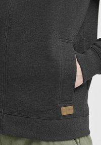 Hoodie zippé gris foncé en tissu doux. Présente des poches latérales et un patch en cuir beige en bas. Poignets et ourlet côtelés.